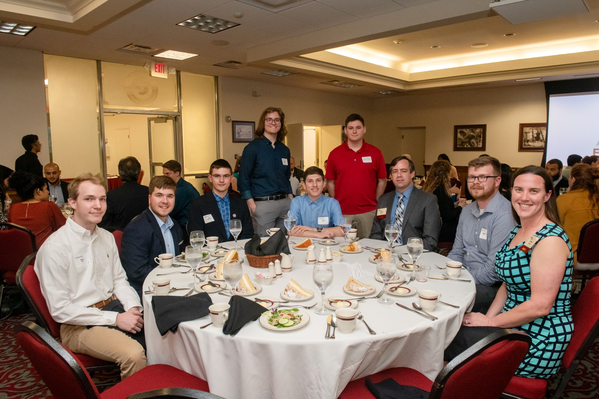 Group of people seated around a table, dressed formally, attending a banquet event with a well-set table featuring plates, cups, and utensils